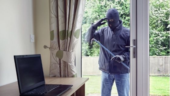 Maskierte Person mit Brecheisen steht vor einer offenen Terrassentür neben einem Laptop