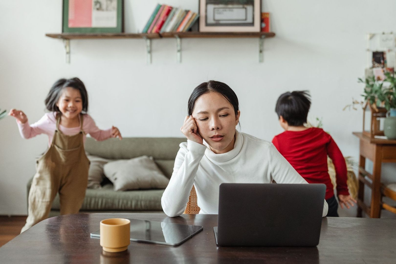 Erschöpfte Mutter am Laptop, während Kinder im Wohnzimmer spielen